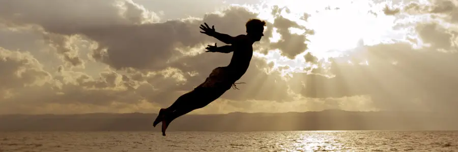Silhouetted figure of a man leaping over water with sun rays and clouds in the background, symbolizing freedom and remote collaboration for SMBs.