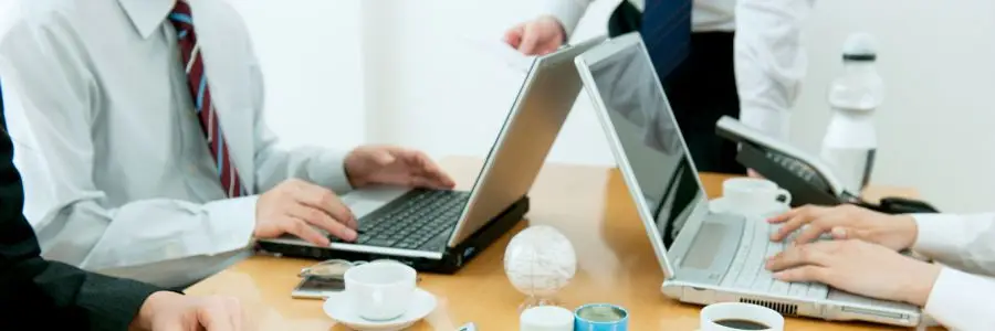 Business meeting with professionals collaborating at a table, using laptops and discussing strategies to enhance office productivity through technology solutions.