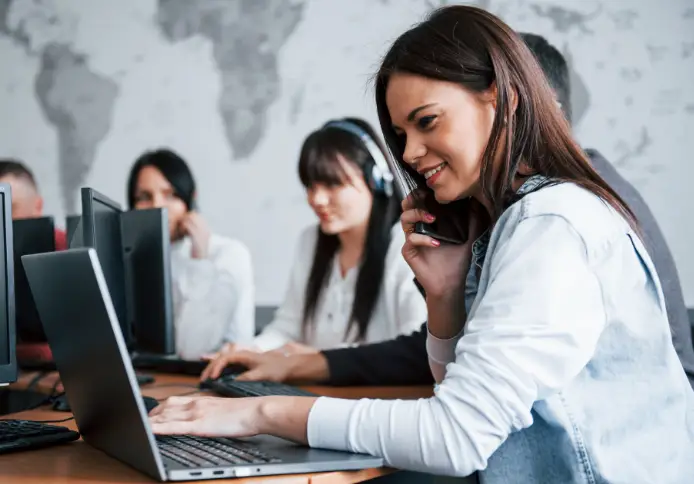 Sign – Managed IT Services, Consulting, and Support for Businesses and Non Profits Woman using laptop and phone in IT support environment, with colleagues working at computers in background, highlighting managed IT services and technology support.